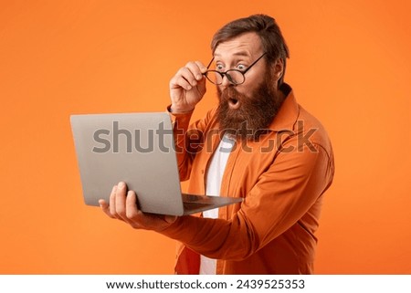 Amazed freelancer guy with red hair and beard looking at laptop computer, touching his glasses in shock, standing with notebook while working online and websurfing on orange studio background Royalty-Free Stock Photo #2439525353