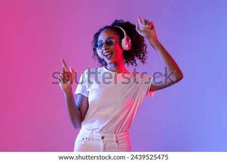 Vibrant young black woman with curly hair enjoying music in wireless headphones and dancing against pink and blue gradient neon backdrop