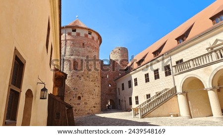 The medieval castle with towers and tiled roofs is built of stone and red brick. The courtyard is paved with stone. The walls have windows and loopholes A staircase with railings leads to the quarters Royalty-Free Stock Photo #2439548169