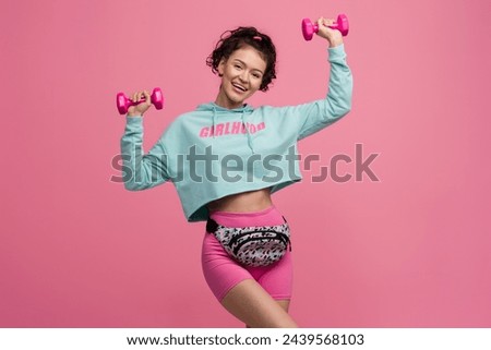 smiling strong slim skinny happy beautiful woman in stylish sport outfit doing workout with dumbbells on pink background isolated in studio, wearing blue hoodie, bag and shorts sportswear Royalty-Free Stock Photo #2439568103