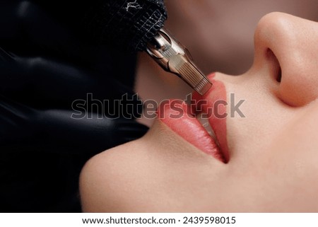 Procedure permanent makeup in beauty salon, tattoo machine with red pigment on woman lips.
