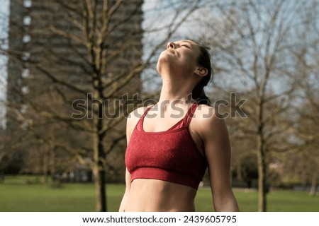 Relaxed image of fitness model meditating in a park with eyes closed. She is wearing a red sport bra and her hair is a ponytail.