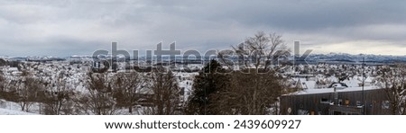 Wide panorama of a snowy urban landscape with distant mountains, blending city life with nature's majesty under a vast cloudy sky.