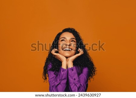 Happy woman with curly hair, wearing casual clothing, smiles with a dreamy expression. Against a orange background, her pensive yet happy expression is vibrant and fun.