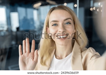 A cheerful young professional woman waving at the camera with a beaming smile in a contemporary office setting. Royalty-Free Stock Photo #2439636659