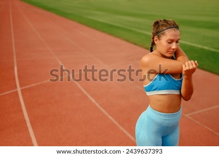 Portrait of a female body conscious athlete standing on running track at stadium and stretching her arms. A fit sportswoman in shape is warming up for cardio training at stadium. Bodycare, healthcare. Royalty-Free Stock Photo #2439637393
