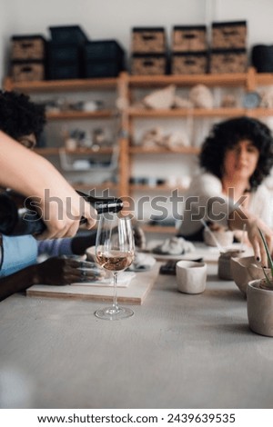 Cropped picture of a hand pouring wine on pottery course with attendees in a blurry background making handcrafts. Close up of hand with wine bottle pouring wine into a glass on pottery class in studio