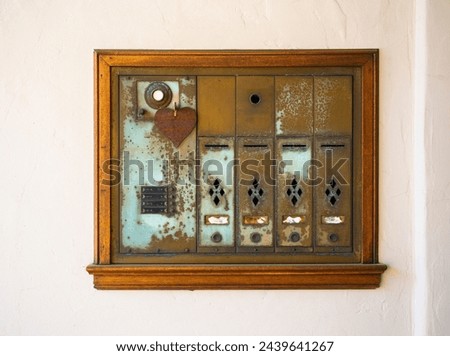 Rusted, corroded, time-worn set of metal mailboxes in small apartment building, with a wood frame and a heart pendant. Gritty and grainy, vintage mood. Against off-white plaster wall. Royalty-Free Stock Photo #2439641267