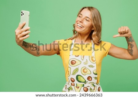 Young housewife housekeeper chef cook baker woman wear apron yellow t-shirt doing selfie shot on mobile cell phone point on herself brag isolated on plain pastel green background. Cooking food concept