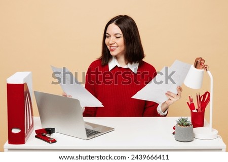Young happy employee business woman wears red sweater shirt sit work at office desk with pc laptop hold paper account documents isolated on plain pastel beige background. Achievement career concept