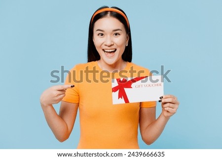 Young woman of Asian ethnicity wear orange t-shirt casual clothes hold point on gift certificate coupon voucher card for store isolated on plain pastel light blue cyan background. Lifestyle concept