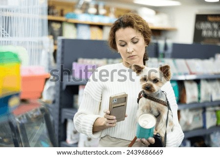 Woman with Yorkshire terrier dog is choosing soft food in tin can for small breed dog, visitor photographing by phone packaging label. Female client scans QR code on feed jar