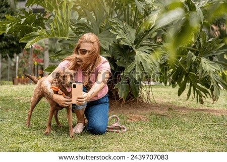A woman squatting with sunglasses taking a selfie with her pet in a park area.