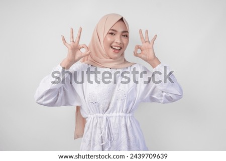 Portrait of Asian Muslim woman in white dress and hijab making an okay, good, nice sign gesture using fingers. Ramadan and Eid Mubarak concept