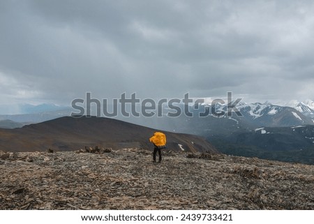 Man in yellow raincoat on stony pass with view to valley against large snow-capped mountain range in gray rainy low clouds. Guy among sharp stones. Big snowy mountains in rain under grey cloudy sky.