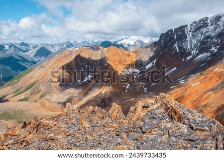 Scenic top view from stony precipice edge to multicolor big rocky ridge in freshly fallen snow and giant snow-capped mountain peak in low clouds. Snow-covered multi-color sharp rocks in huge mountains Royalty-Free Stock Photo #2439733435