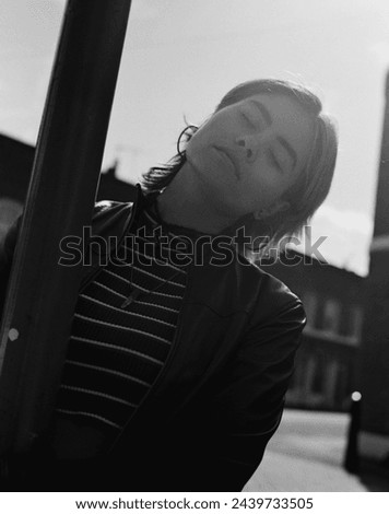 Analog image of young woman posing around a street light. Her eyes are closed and she is wearing a leather jacket. Back-light. Analog film image made with medium format camera.
