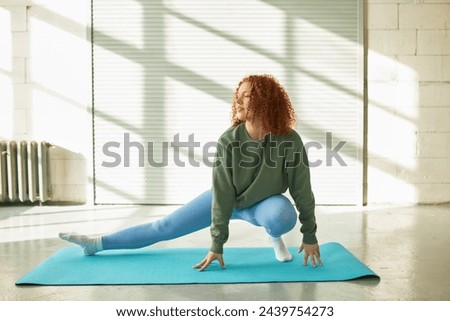 Full-length portrait of flexible redhead woman in sports clothes at gym stretching leg, bending one knee, training indoors, keeping fit and losing weight, enjoying physical exercises Royalty-Free Stock Photo #2439754273