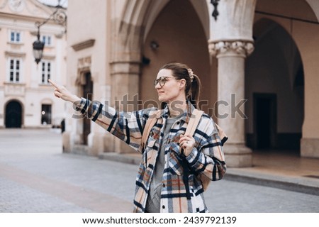 Young happy woman traveler with a backpack traveling Europe in autumn. Stylish female foreigner examines architectural monument during her long-awaited vacation Royalty-Free Stock Photo #2439792139