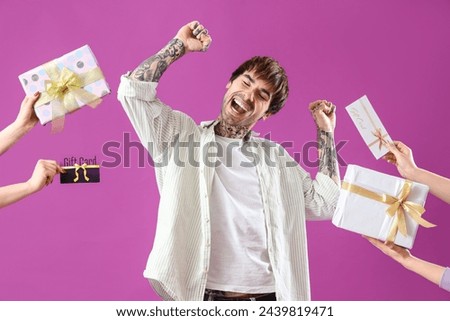 Happy young tattooed man with gift cards and presents on purple background