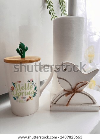 a mug with the inscription hello Spring and a lid with a cactus next to a wooden towel holder and a bird figurine.