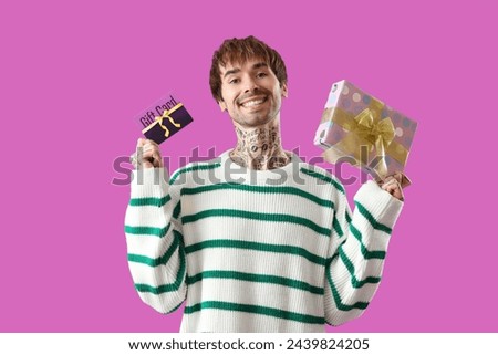 Young tattooed man with gift card and present on purple background
