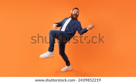 Energetic guy with red beard enjoying music through headphones while playing invisible guitar, having fun over orange studio backdrop. Full length shot, panorama with copy space