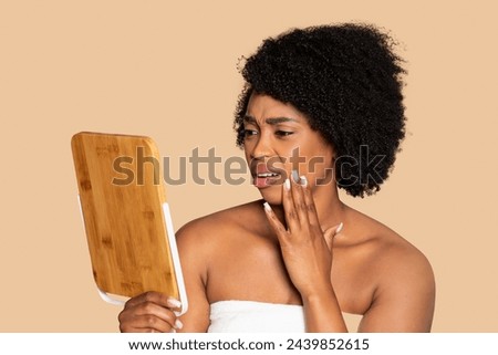 Concerned black woman in towel, inspecting her complexion with wooden mirror, expressing skin health worries, on harmonious beige background Royalty-Free Stock Photo #2439852615