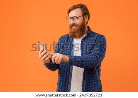 Casual European guy with red hair and beard browses internet on his smartphone, wearing eyeglasses while texting and websurfing over orange studio background. Concept of mobile app Royalty-Free Stock Photo #2439852721