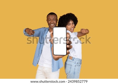 Excited millennial glad African American couple presenting a smartphone with a blank screen towards the camera, ideal for app or website promotion, on a cheerful yellow background Royalty-Free Stock Photo #2439852779