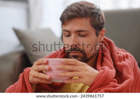 autumn and winter season of sneezes. Young Caucasian man with flu on sofa at home. Young Caucasian man with flu feels cold in living room at home drink hot tea in cup.