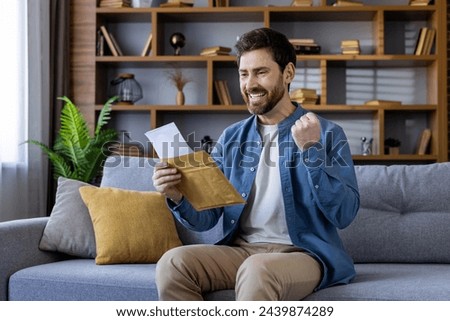 A joyful man sitting on the sofa at home, holds an envelope with excitement, showcasing a positive reaction with a clenched fist.