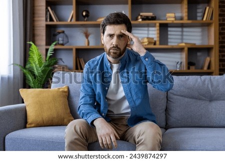 A pensive adult male with a beard sits on a sofa, appearing stressed or troubled, surrounded by a cozy home environment. Royalty-Free Stock Photo #2439874297