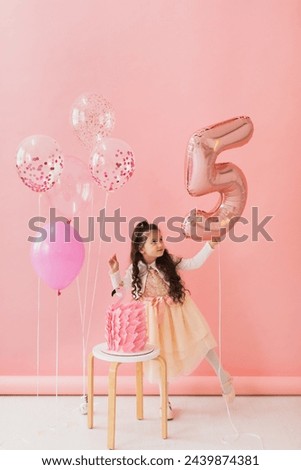 Adorable little girl in a pink dress posing next to a flamingo birthday cake and celebrating her fifth birthday on a pink background