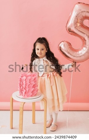 Adorable little girl in a pink dress posing next to a flamingo birthday cake and celebrating her fifth birthday on a pink background