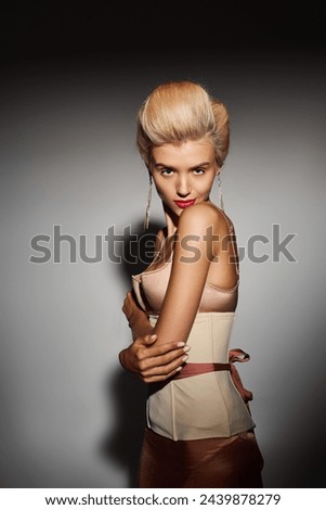 blonde girl in corset dress with red lips hugs herself and looks defiantly on grey background Royalty-Free Stock Photo #2439878279