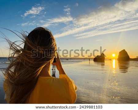 Back view of a young woman taking a picture of a sunset at the beach with mobile phone. Praia dos Tres Irmaos, Algarve Portugal.