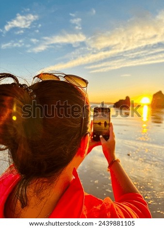 Back view of a young woman taking a picture of a sunset at the beach with mobile phone. Praia dos Tres Irmaos, Algarve Portugal.