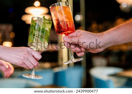Two hands clink cocktail glasses in a modern bar setting, celebrating. One glass holds green drink with mint leaves and lime slices, possibly a mojito. other contains a red-orange fruit-based cocktail