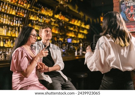 Young guy attracts attention of brunette girl with long hair in blouse. Friends sit with cocktails at bar near trendy picture on wall