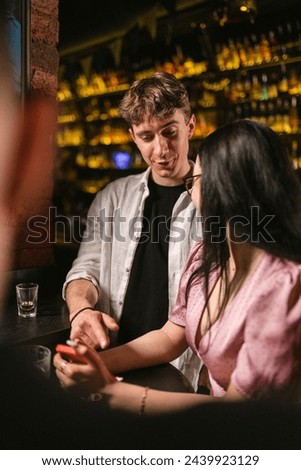 Brunette lady with eyeglasses communicates with companion near bar table. Young people speak about mobile phone after party