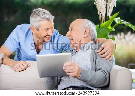 Laughing male caretaker and senior man using tablet computer at nursing home porch