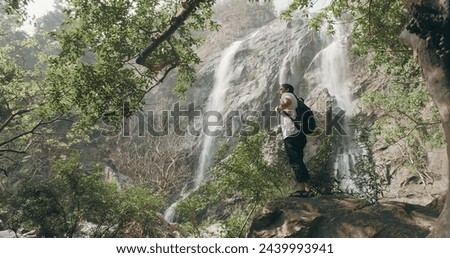 A man is standing on a rock near a waterfall. He is wearing a backpack and a white shirt. The scene is peaceful and serene, with the sound of the waterfall in the background