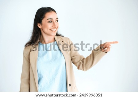 Joyful pretty indian or arabian brunette woman, dressed in elegant suit, pointing with finger to the side at empty space, standing on isolated white background, looks away, smiling happily
