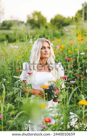 Beautiful Blonde Model Sitting in Zinnia Wildflower Garden, Colorful Golden Hour Sunset Bridal Portrait of Bridal Model with Loose Hair Wearing Dress with Puffy Sleeves
