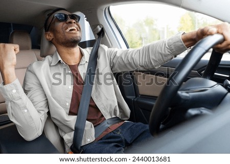 Joyful African American man in green glasses dances in the car, sings while driving. Road fun Royalty-Free Stock Photo #2440013681