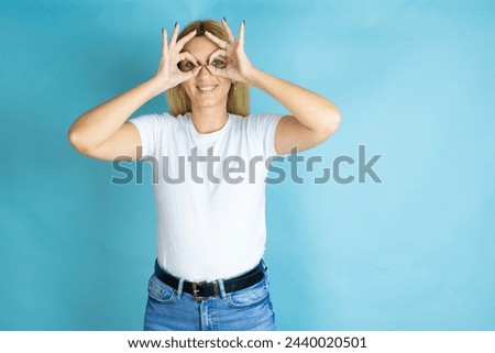 Young beautiful woman wearing casual t-shirt over isolated blue background doing ok gesture shocked with smiling face, eye looking through fingers