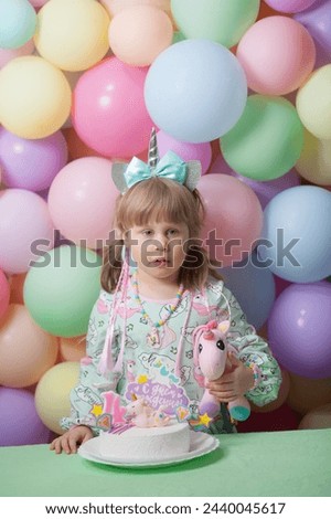 A little girl in a unicorn dress and hairband is holding a pink unicorn soft toy by her birthday cake. Four years-old celebration. Pastel latex balloons background. Vertical image. 
