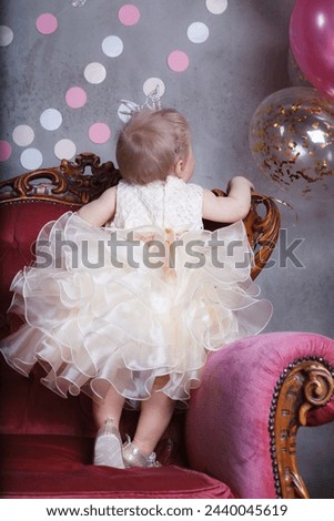 A beautiful one-year-old baby girl in a festive dress with a crown on her head is standing on a pink chair. Back view. Vertical image. 