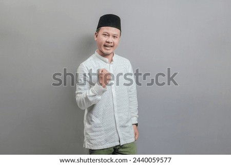 Happy asian muslim man fun successful raised fist hands wearing a koko shirt and peci with shades of the fasting month, isolated on a gray background